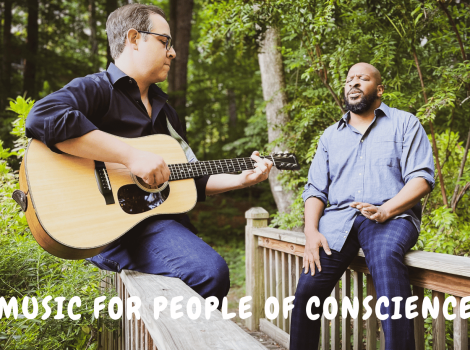 Rabbi Lapidus playing guitar next to Myles as they sit outdoors on a bridge