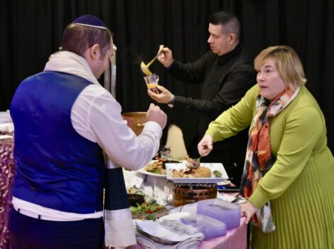 man in blue sweater vest samples food being served by woman in green dress at the Simcha Fair