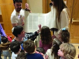 adults hold up an open torah scroll in front of a group of young children