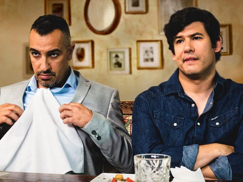 movie still from Maktub with 2 men. On left has a napkin tucked into his shirt, right is smiling in a blue shirt